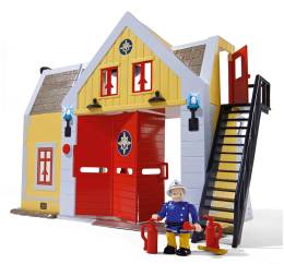Brandman Sams brandstation syns framifrån. Gul byggnad med stor röd port. Svart trappa går upp längs med sidan av huset till en öppen röd dörr. Framför stationen står en man med brandutrustning och grå mustasch.